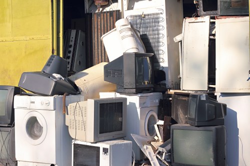 A large pile of discarded household appliances and electronic waste is stacked outdoors, including several washing machines with circular doors, old computer monitors with glass screens and plastic casings, a microwave oven, and various metal cabinets and drawers. The appliances are arranged haphazardly, some leaning against each other and others resting on the ground or on top of each other. The scene appears to be in a dedicated waste collection or rubbish storage area, possibly part of a rubbish removal service in the Nottingham or nearby postcode area, as suggested by the context of Garden Clearance Mottingham. The environment is an industrial-looking outdoor space with visible metal storage units, and the lighting indicates daylight, highlighting the different textures and finishes of the varied objects, predominantly in white, black, and metallic tones, with some signs of weathering or wear. This image illustrates typical electronic waste that a rubbish removal service might handle from domestic or commercial properties in the Nottingham region.
