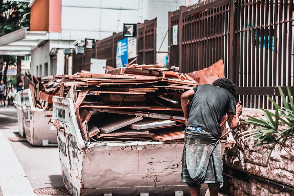 A man with curly black hair wearing a dark t-shirt and plaid shorts stands on a sidewalk next to a large, white skip filled with a variety of discarded roofing materials, including terracotta roof til